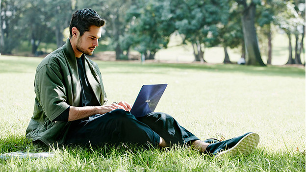 Man sitting in the grass works on Asus laptop in park.