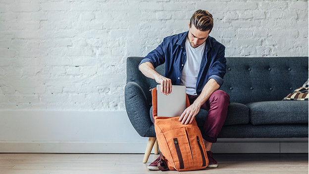 Man on couch puts laptop in orange backpack.