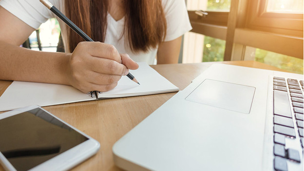 Frau schreibt in ein Notizbuch, während sie an einem Notebook auf ihrem Schreibtisch arbeitet.