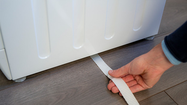 Führe den Gurt genau mittig unter der Waschmaschine hindurch.